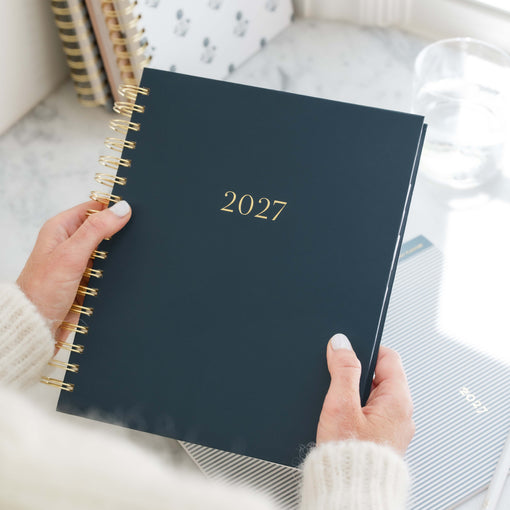A person holding a navy blue 2027 spiral-bound planner with gold lettering and gold wire binding, set against a bright, marble-topped background with other stationery.