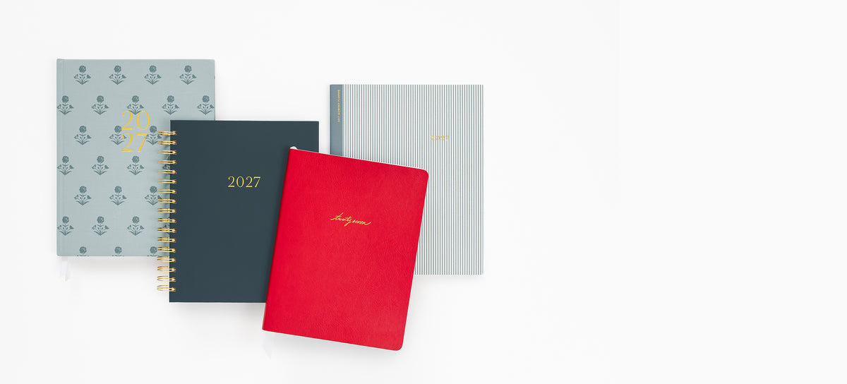 Planners arranged on a white background: a light blue patterned planner, a dark navy 2027 spiral planner, and a red notebook with gold script.