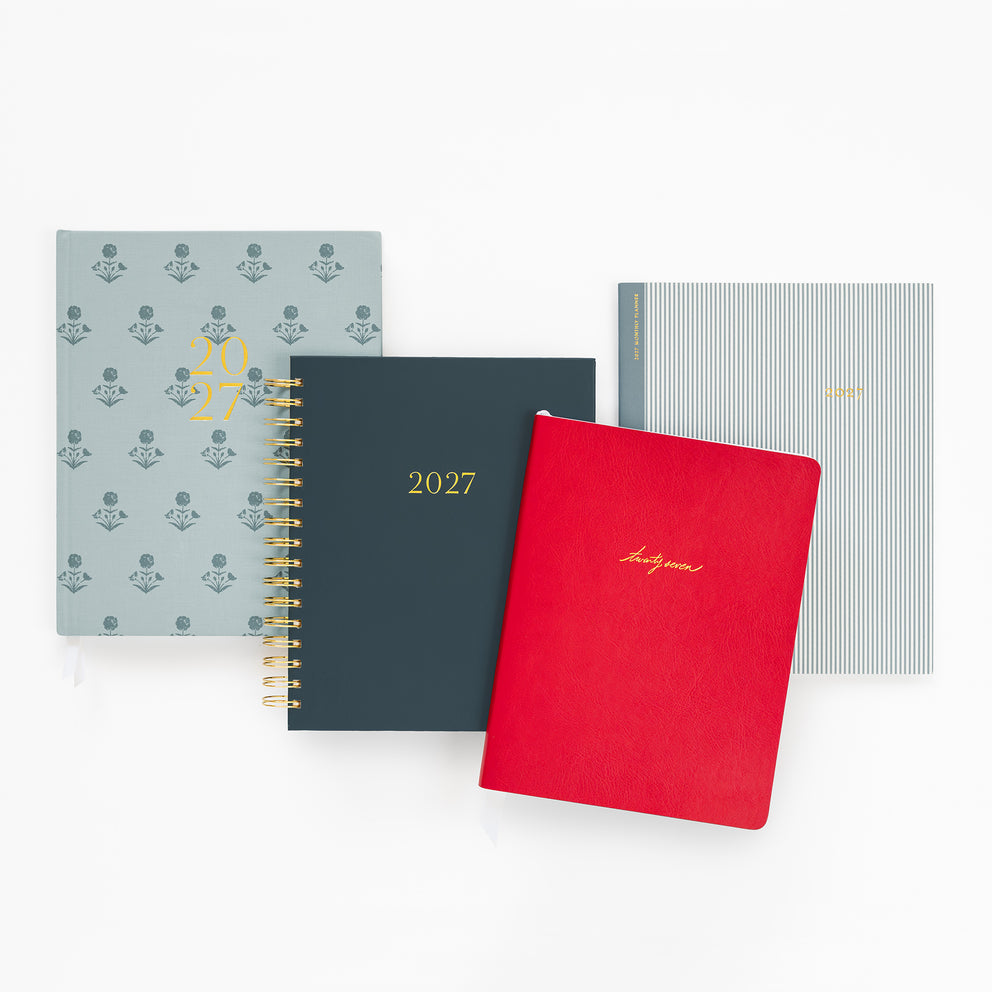 Four 2027 annual planners arranged in a fan-like stack on a white background. The set includes a light blue planner with a repeating floral pattern and gold '2027' text, a dark slate blue spiral-bound planner with gold '2027' text, a bright red hardcover planner with gold 'Happy Birthday' script, and a light blue-gray striped planner with gold '2027' text.