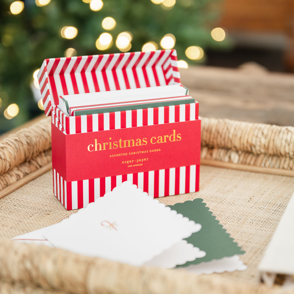 A red and white striped box labeled 'christmas cards' sits on a woven tray with a few sample cards in front, against a softly blurred holiday background with warm lights from the christmas tree
