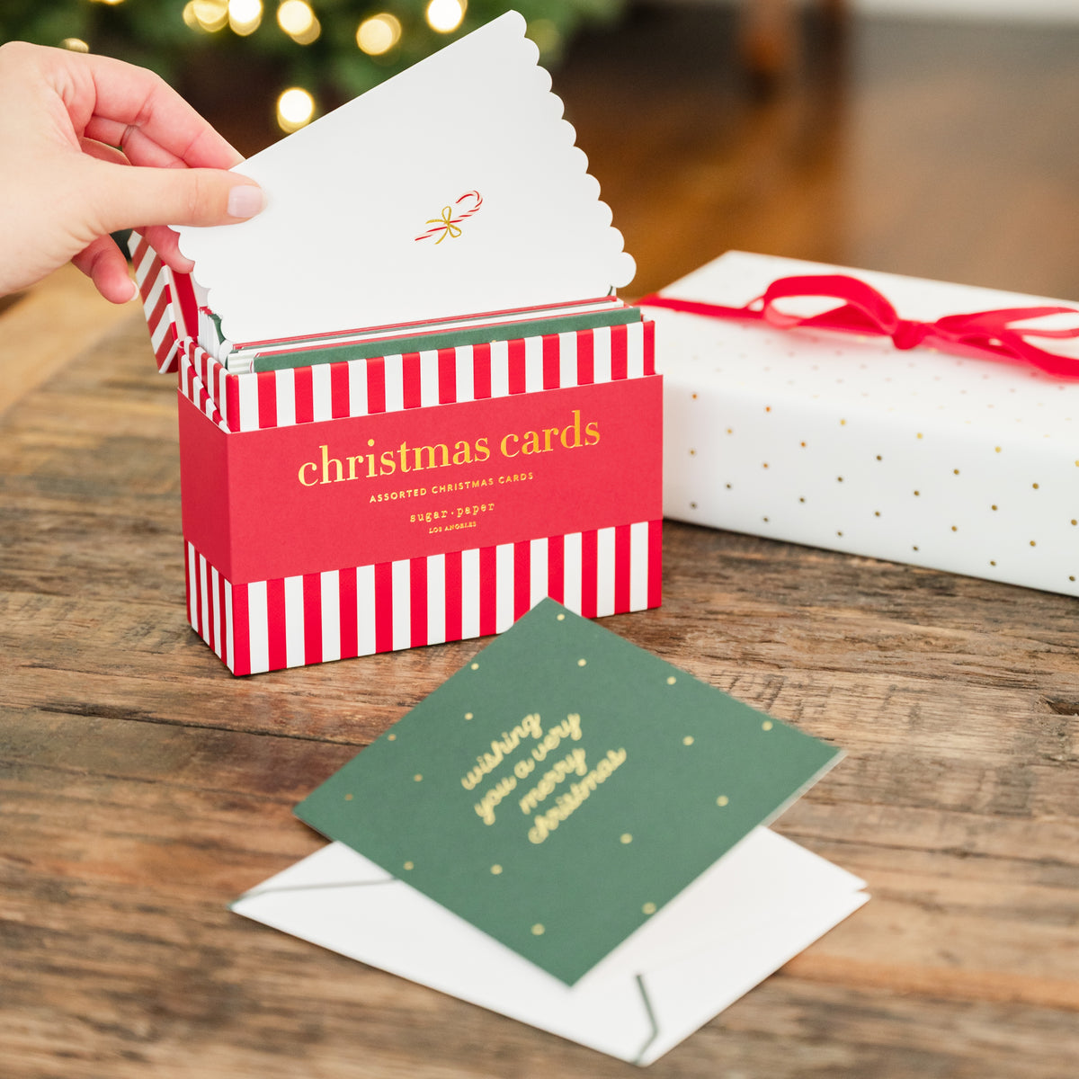 A hand is opening a red and white striped box of Christmas cards on a wooden table. A green card with gold text printed with 'wishing you a merry christmas' is pulled out and placed on top of a white envelope. There is a christmas tree and holiday gift in the background