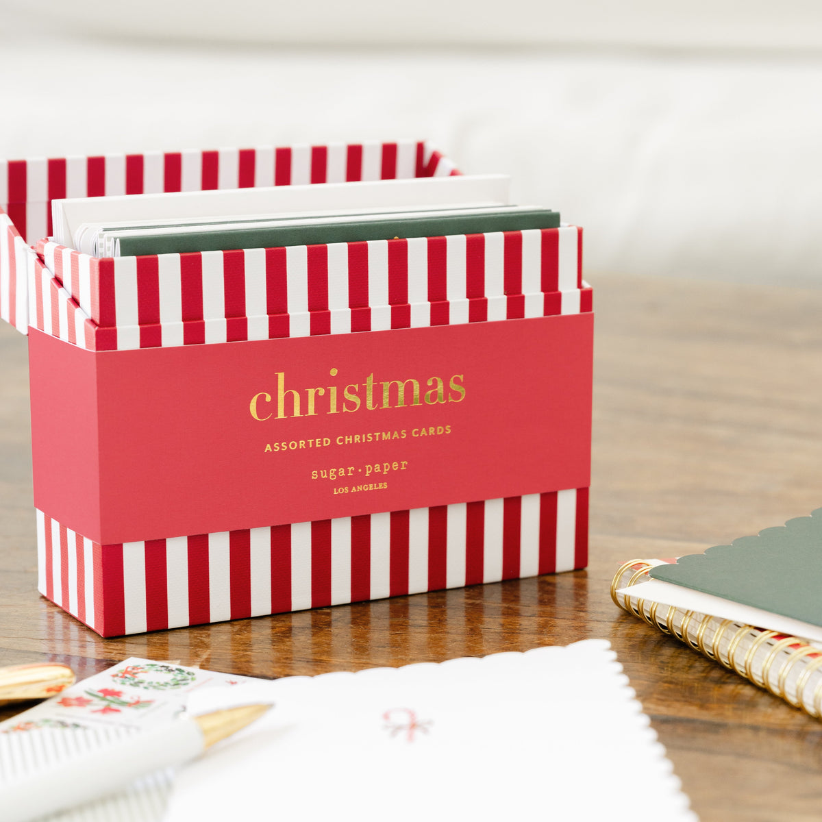 A red and white striped gift box labeled 'christmas' with gold lettering sits on a wooden table, surrounded by white and green cards and holiday stamps