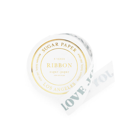 A roll of white sugar paper ribbon with blue 'peace love joy' text printed on it, shown on a clean white background. The round label on the roll reads 'SUGAR PAPER RIBBON LOS ANGELES' in gold foil