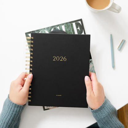 Person holding a black planner with '2026' on it, placed on a white surface with a cup of coffee and magazine
