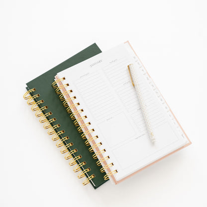 Two small spiral-bound planners, one green and one blush open to monthly goal page with gold spirals