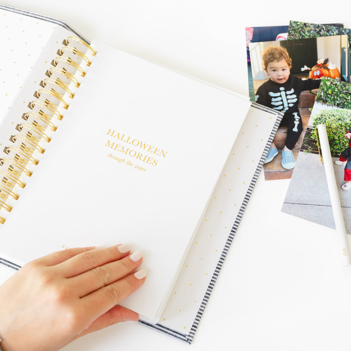 An open spiral-bound photo album with gold accents and the words 'HALLOWEEN MEMORIES through the years' on the cover, held by a hand over a white surface with printed Halloween photos nearby.