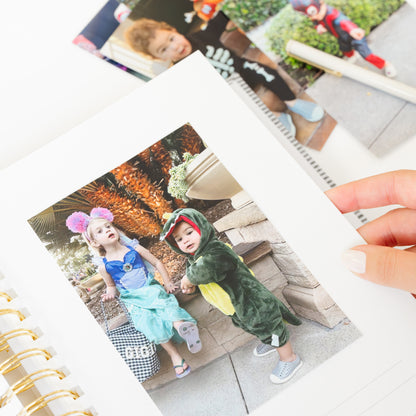 A hand holds a printed photo page showing two children in Halloween costumes in a gold spiral bound book with white pages, with other printed photos and a pen in the background