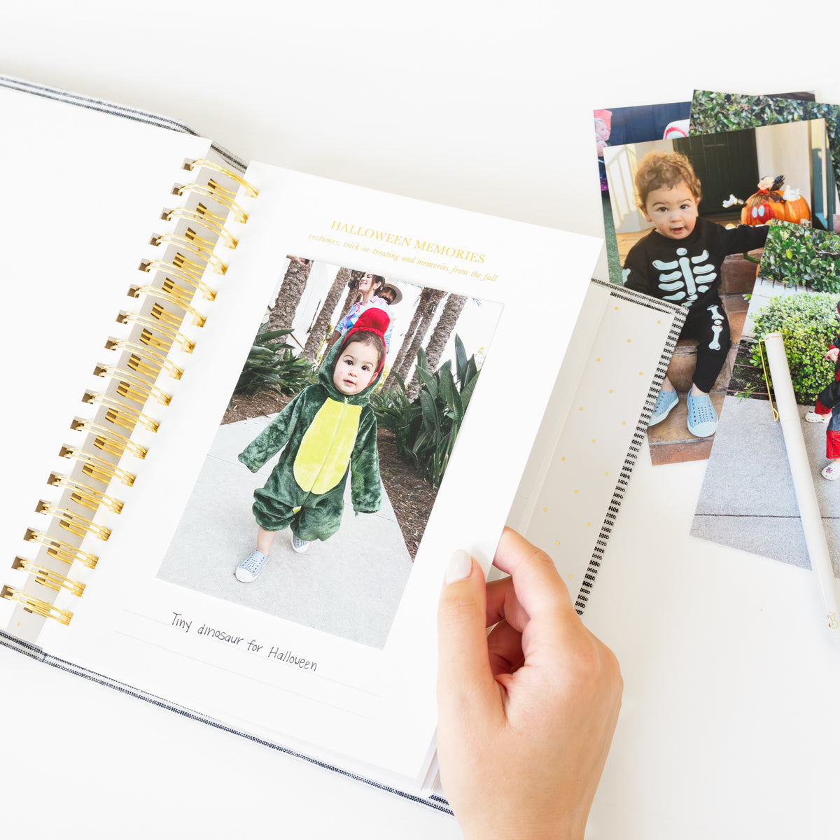 An open spiral-bound photo album on a white surface, showing a printed photo of a child in a green dinosaur Halloween costume. A hand is holding the page, and several printed Halloween photos are stacked to the right.