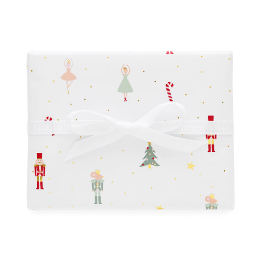 A square gift box wrapped in white wrapping paper with a festive Christmas print featuring nutcrackers, ballerinas, candy canes, a Christmas tree, and gold dots, tied with a white ribbon