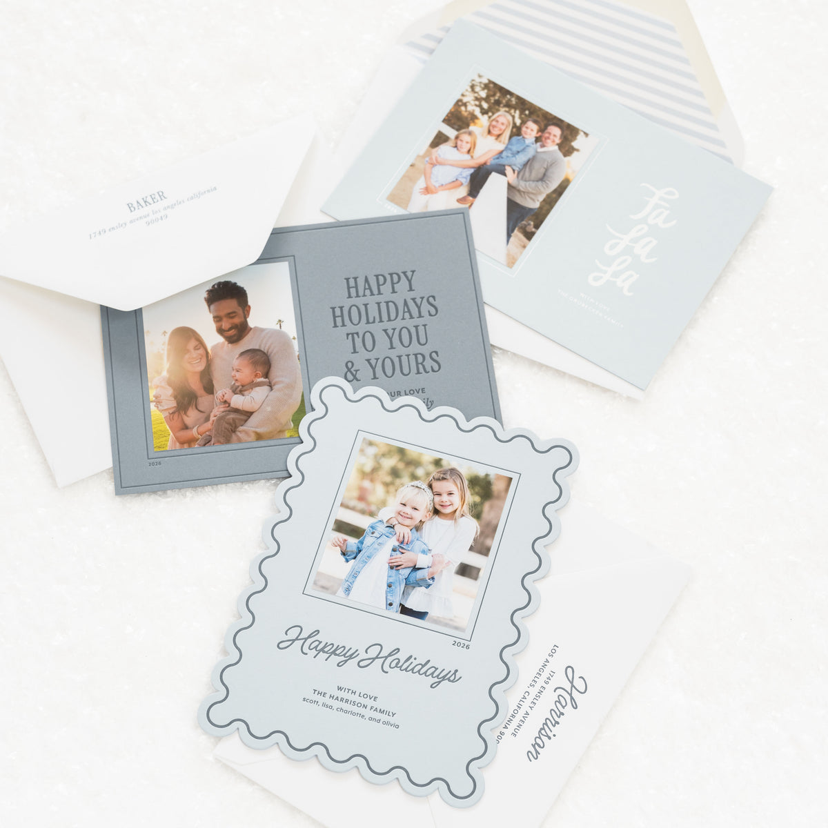 A styled flat-lay of holiday greeting cards and envelopes on a white textured surface. The cards feature light blue, white, and gray designs with sample family photographs and elegant script text including 'Happy Holidays' and 'Let Love In'.