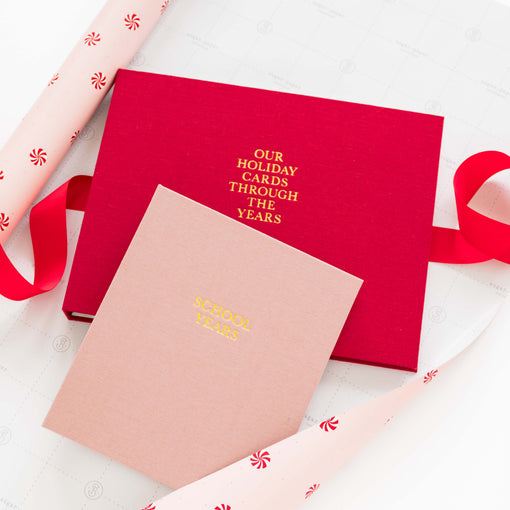 A red box labeled 'OUR HOLIDAY CARDS THROUGH THE YEARS' in gold foil sits on a white surface with a matching pink book reading 'SCHOOL YEARS' in gold foil. Red ribbon and pink wrapping paper with red candy motifs are arranged around the items.