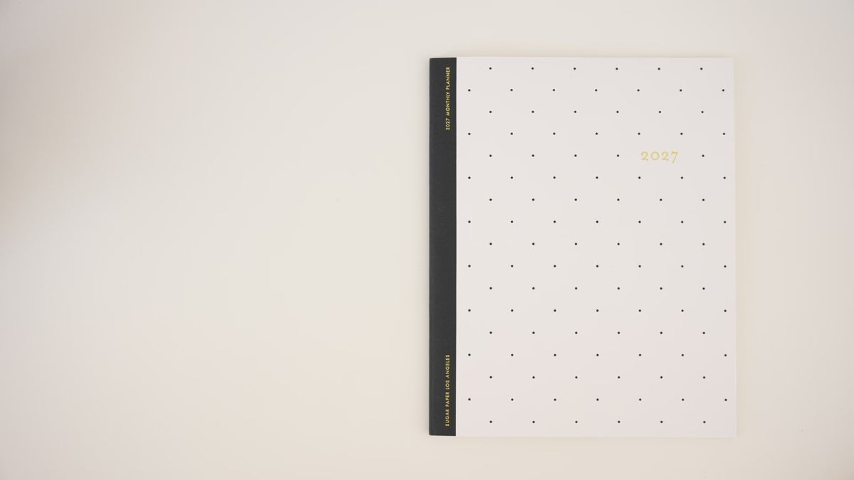 Load video: A video of someone opening the flat monthly 2027 planner with a white with gold dot pattern and black tape binding printed with '2027'