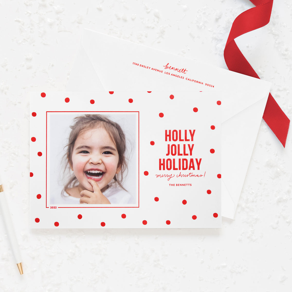White holiday photo card printed with red dot pattern and 'holly jolly holiday merry christmas' in red ink paired with a white envelope with red return address