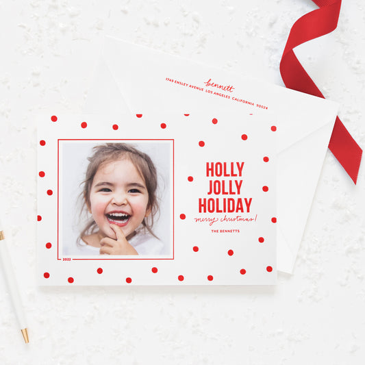 White holiday photo card printed with red dot pattern and 'holly jolly holiday merry christmas' in red ink paired with a white envelope with red return address